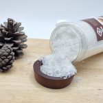 نمک سفید رنگ بیرون ریخته شده از بسته بندی نمک حمام در ظرف چوبی با عصاره نارگیل به همراه میوه کاج در کادر